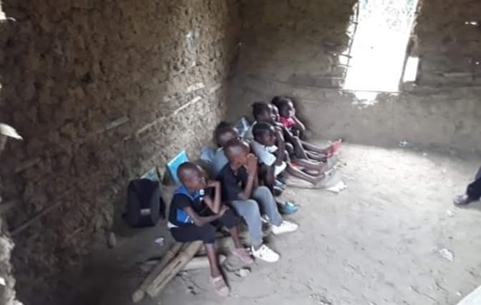 Students sit on makeshift bench of sticks to take lesson